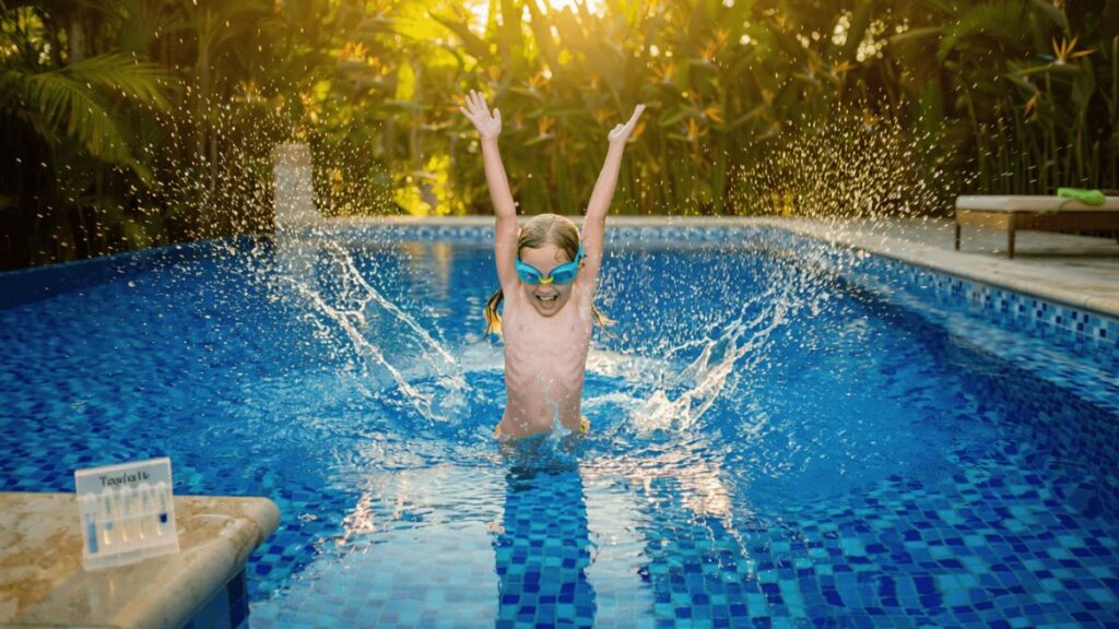 Young child aged 6 to 8 playing joyfully in a crystal clear backyard swimming pool with perfectly balanced pH, splashing clean blue water in warm afternoon sunlight