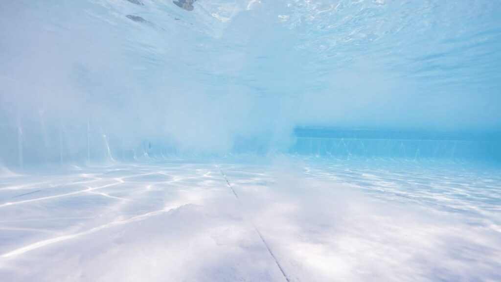 Close-up of new white pool plaster surface with fine white calcium carbonate dust settling on pool floor and suspended in water, causing pH to rise during curing process