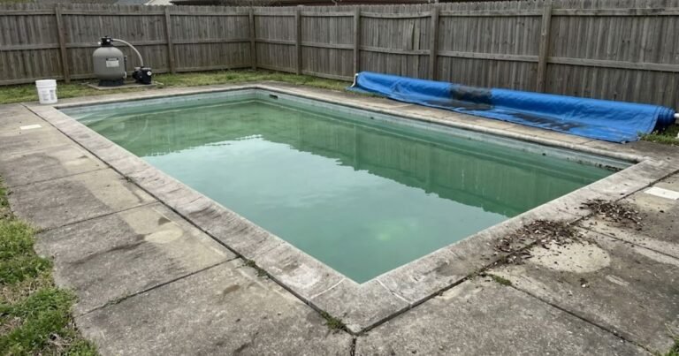 Folded blue winter pool cover leaning against fence next to a residential in-ground concrete pool with murky water at season opening