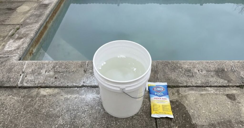 White 5-gallon bucket with pre-dissolved pool shock granules sitting on concrete pool deck beside an in-ground pool during spring opening
